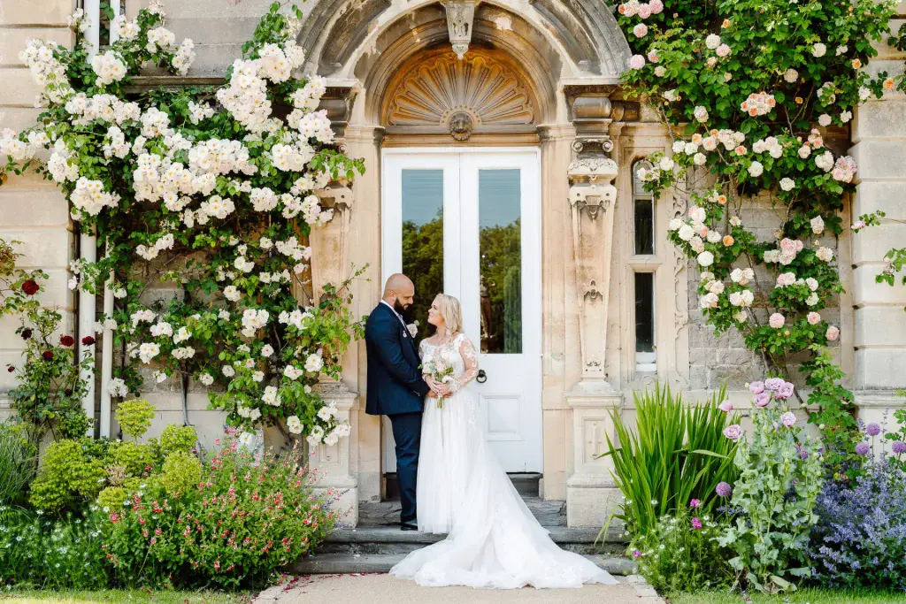bride and groom stand looking at one another underneath a wall of blooming roses at clevedon hall in somerset