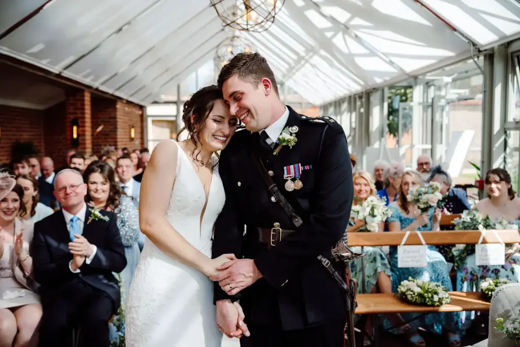 bride and her husband dressed in ceremonial military uniform huddle in their wedding ceremony inside the glasshouse at Syrencot wedding venue during their wedding live stream