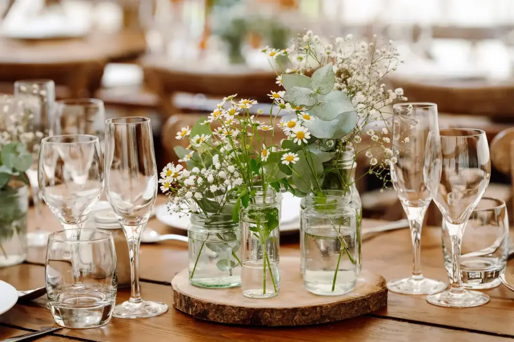 small daisies in glass jars on a wooden place holder at a wedding breakfast illustrating a DIY wedding flowers arrangement