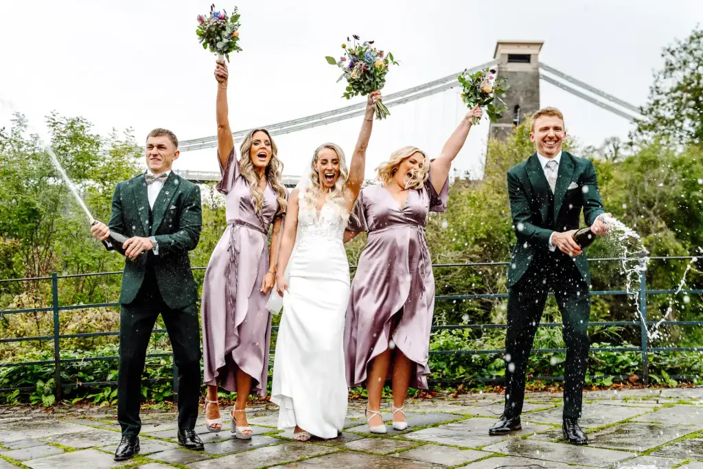 a group of people posing for a picture while spraying champagne in front of the Bristol suspension bridge in Clifton