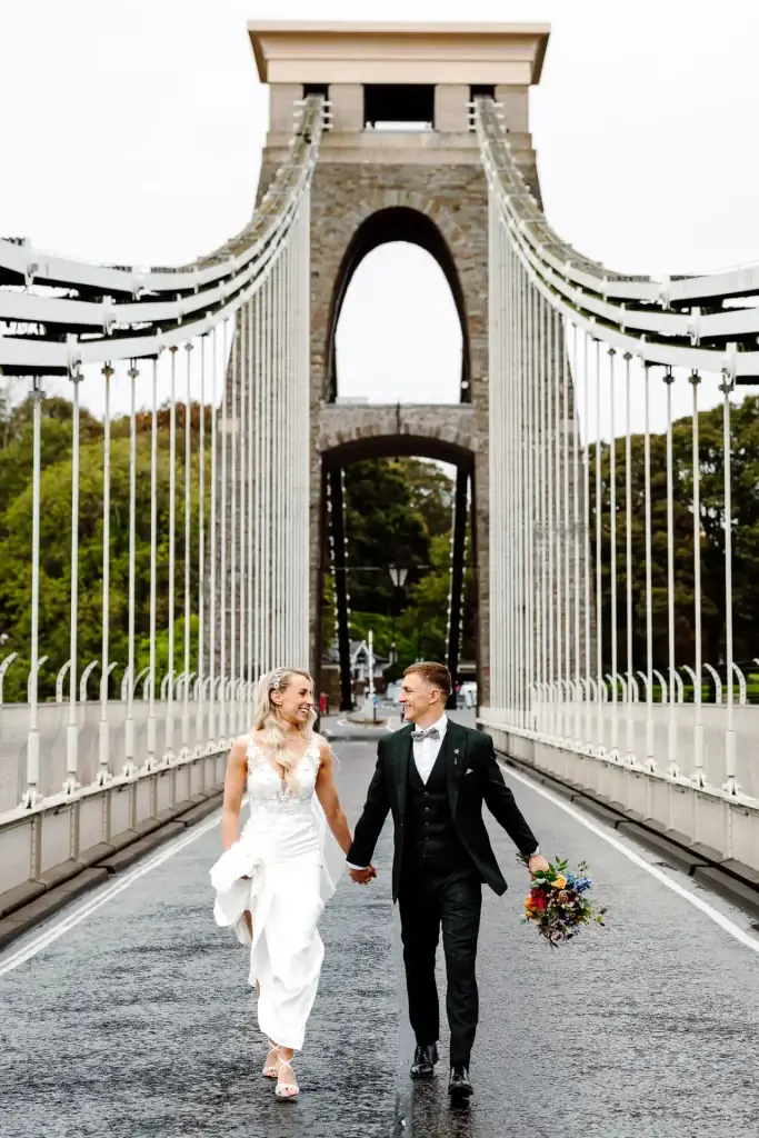 bride and groom walk down the middle of the clifton suspension bridge looking at one another in this example of bristol wedding videography