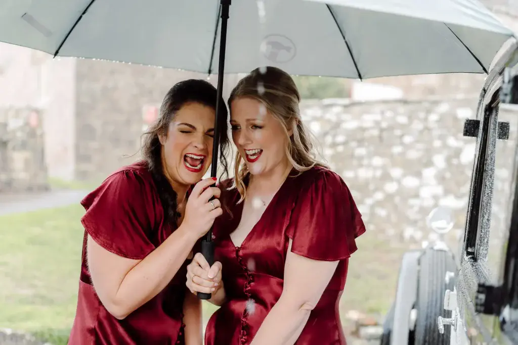 two bridesmaids in red dresses take cover under an umbrella on a rainy wedding day and laugh at the situation